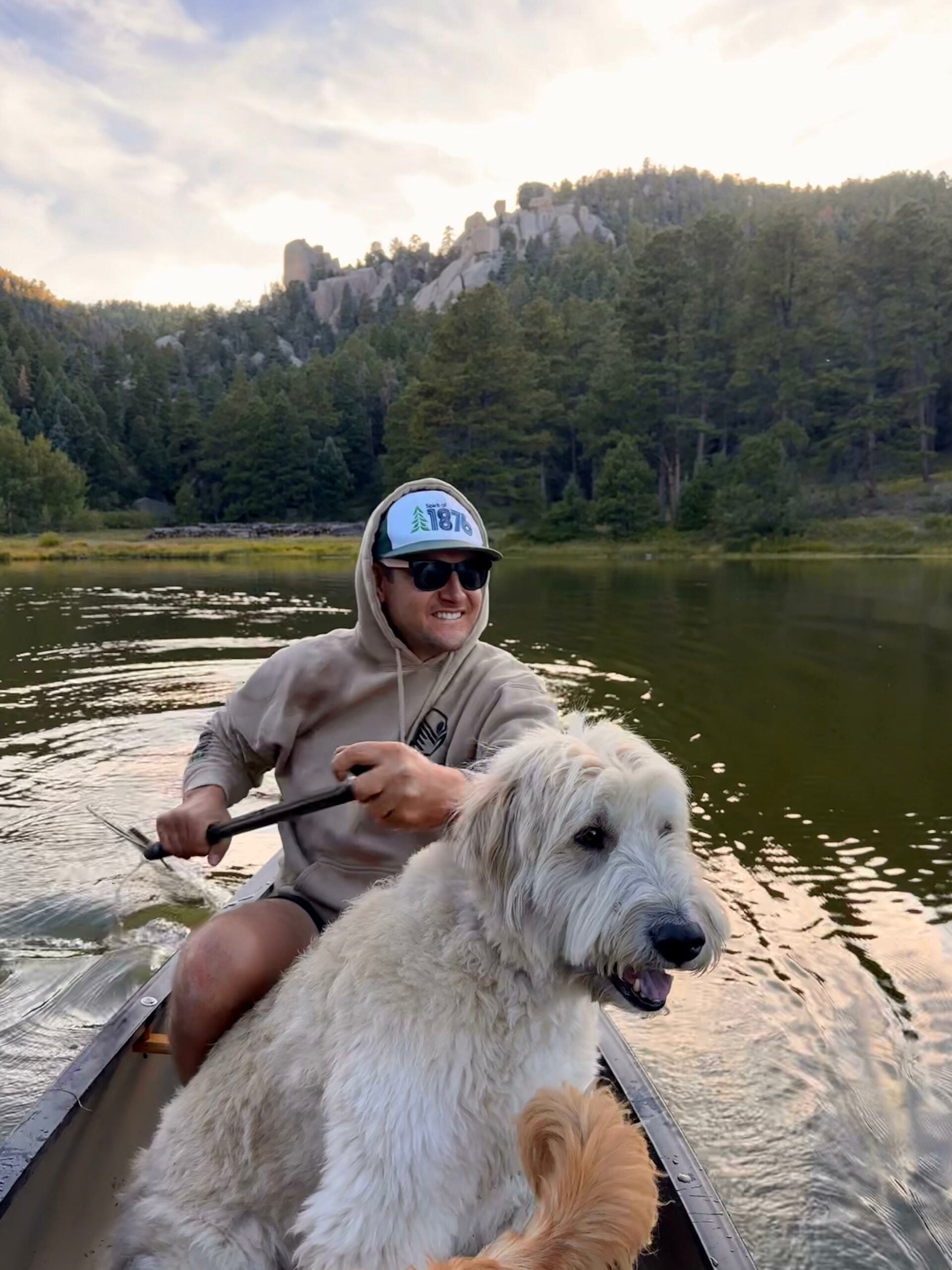 Man in a kayak with a large white dog on a lake surrounded by trees and mountains.